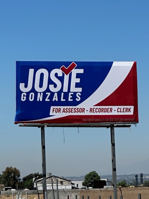 A billboard features a political advertisement with a red, white, and blue color scheme. It reads "Josie Gonzales for Assessor - Recorder - Clerk" with a checkmark integrated into the name. Trees and buildings are visible in the background.